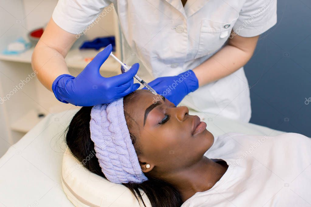 Close up of beautician expert's hands injecting botox in female forehead og young African girl.