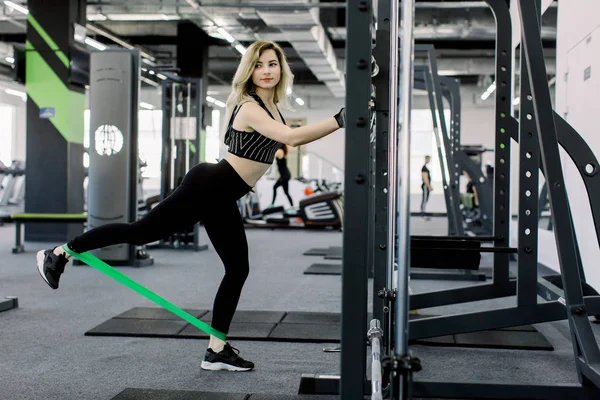 Jeune femme blonde sportive fait des exercices sur les jambes avec des élastiques de fitness sportif. Photo de la femme musclée en entraînement de vêtements de sport dans la salle de gym. Force et motivation . — Photo