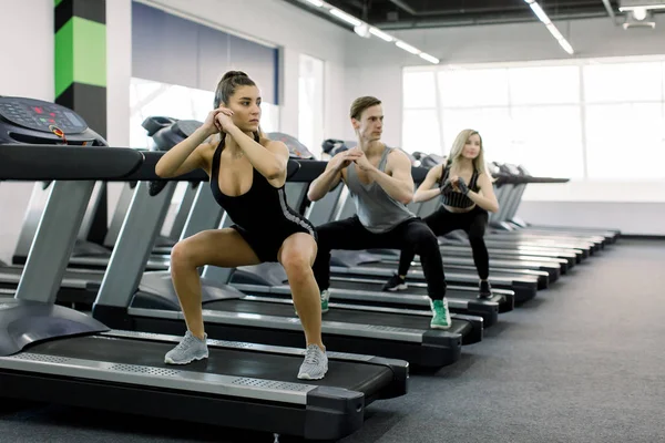 Young active people, two girls and boy, doing squats while standing on a treadmill. Group of fit people doing sport in the gym.