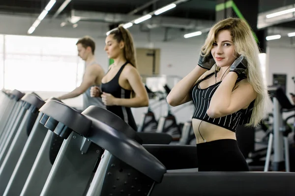 Jovens correndo em uma esteira no health club. Menina correndo na esteira e ouvir música no ginásio, juntamente com amigos — Fotografia de Stock