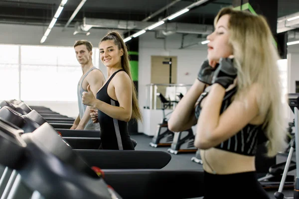 Jovens amigos esportivos fazendo exercícios cardiovasculares em esteira rolante estacionária no ginásio. Duas meninas e seu treinador bonito correndo na esteira da máquina no clube de ginásio de fitness — Fotografia de Stock