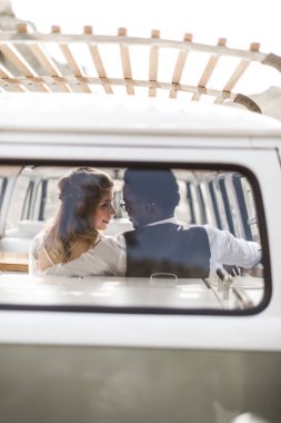 Gezi, aşk ve düğün konsepti sırasında rahatlatıcı. Neşeli genç çok ırklı çift birbirlerine bakarak ve retro minivan arka koltuklarda otururken gülümseyen geri görünümü