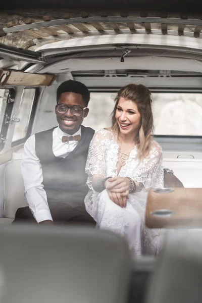Hippie miłość. Szczęśliwa Młoda para, afrykański mężczyzna i kaukaski kobieta klejenia się do siebie i uśmiechnięty siedząc na tylnym siedzeniach swojego minivana, świętując ich ślub — Zdjęcie stockowe