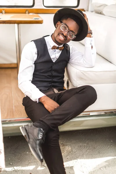 Retrato de cerca del hombre africano guapo en traje elegante y sombrero sonriendo mientras está sentado en minivan de autobús retro en el cañón —  Fotos de Stock