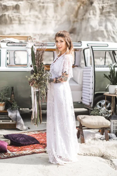 summer, canyon, desert, wedding concept - smiling young hippie women in white long dress, holding rustic flower bouquet, standing in front of green retro minivan car in canyon