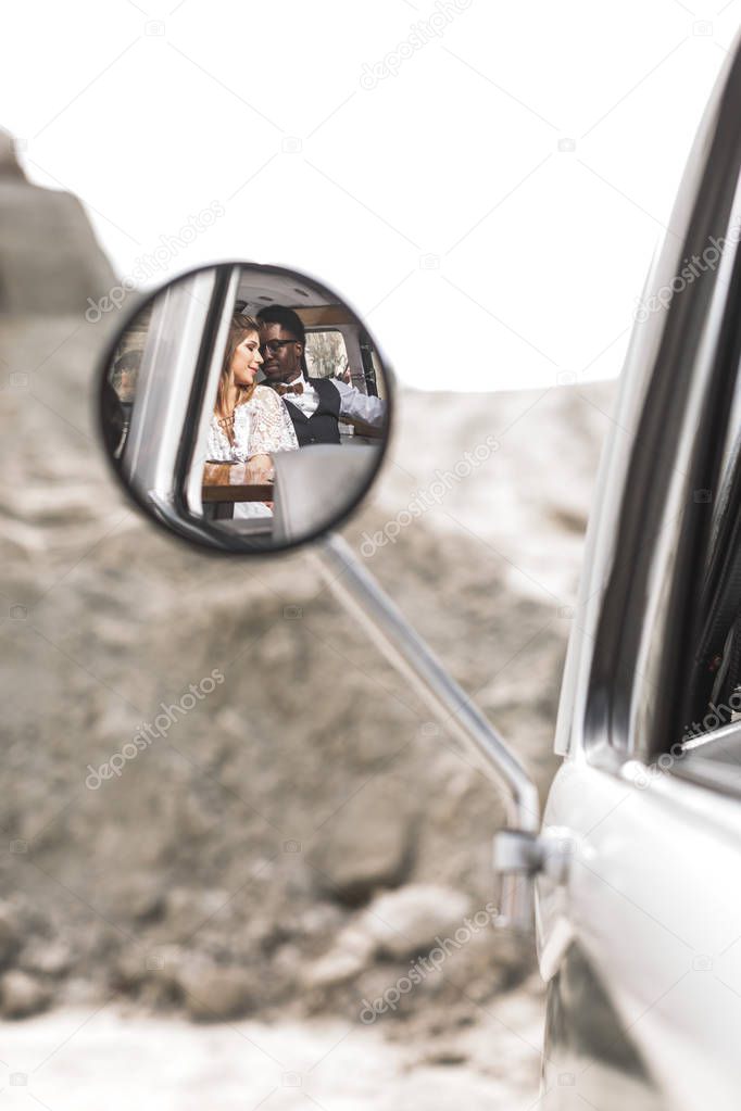Photo of the reflection in the mirror of young wedding hippy couple man and woman smiling and hugging each other while sitting in retro minivan in canyon