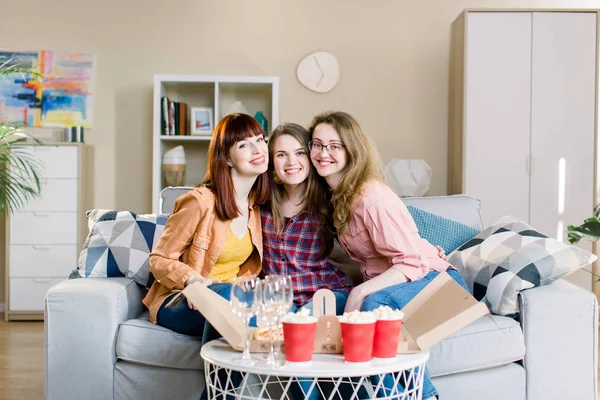 Portrait of three happy young female friends having home party with pizza, popcorn and wine, sitting on the gray sofa in dining room at home. Pizza party, female friendship