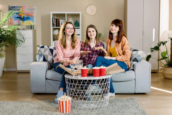 friendship, people, pajama party and junk food concept - happy young three women or girls eating pizza, popcorn and drinking wine at home