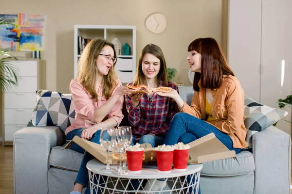 friendship, people, pajama party and junk food concept - happy young three women or girls eating pizza, popcorn and drinking wine at home