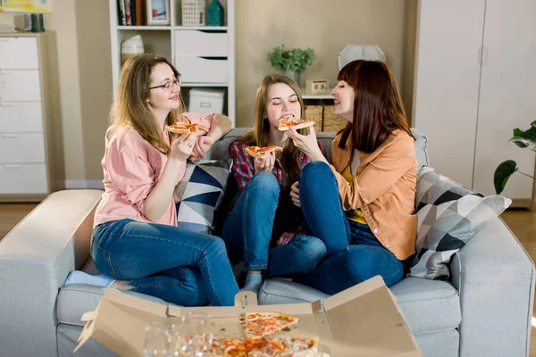Mooie jonge meisjes in jeans en rokken die pizza eten, praten en glimlachen thuis. Vrienden eten pizza, vriendschap, mensen, pyjama party, entertainment en junk food concept — Stockfoto
