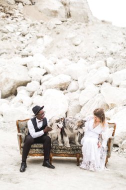 Şık hippiler damat ve gelin büyük bir köpek ile çölün ortasında bir kanepeüzerinde oturur. Başarılı ve stil kavramı