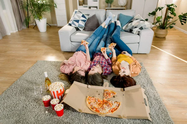 Three Girlfriends in jeans and shirts eating Pizza at home, lying on the floor and put legs on the sofa — Stock Photo, Image