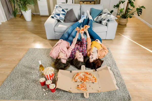 Bir grup genç bayan arkadaş pizza yiyor. Üç kız gri kanepede bacaklarıyla yerde yatarken pizza yiyor. Ev partisi. — Stok fotoğraf