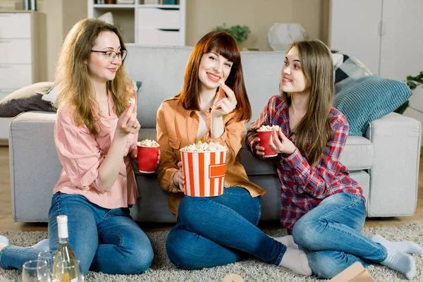 Amistad de mujeres, fiesta en casa. Tres hermosas amigas divertidas con palomitas de maíz en las manos sentadas en el suelo en una acogedora habitación en casa y mirando a la cámara —  Fotos de Stock