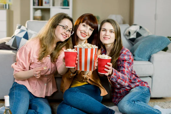 Women friendship, home party. Three beautiful funny young girl friends with popcorn in hands sitting on the floor in cozy room at home and looking on camera