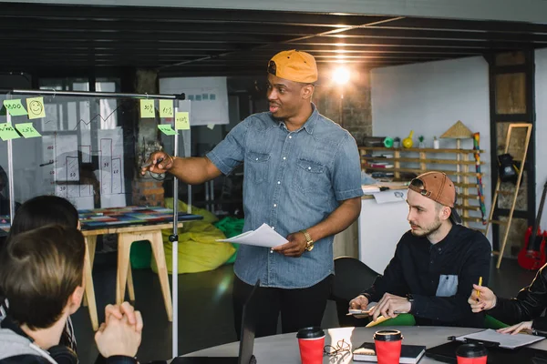 Full concentration at work. Corporate team working colleagues working in modern office. African man making presentation near the glass office wall and showing results of the analysis