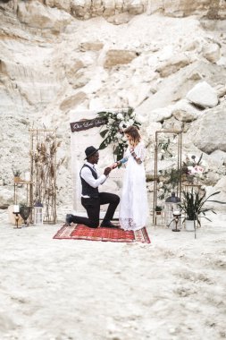 Boho tarzında giyinmiş düğün çifti, düğün kemerinden önce el ele tutuşarak kalıyor. Afrikalı damat dizinin üzerinde duruyor ve Kafkas gelini için alyans takıyor..