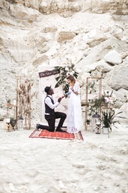 Boho tarzında giyinmiş düğün çifti, düğün kemerinden önce el ele tutuşarak kalıyor. Afrikalı damat dizinin üzerinde duruyor ve Kafkas gelini için alyans takıyor..