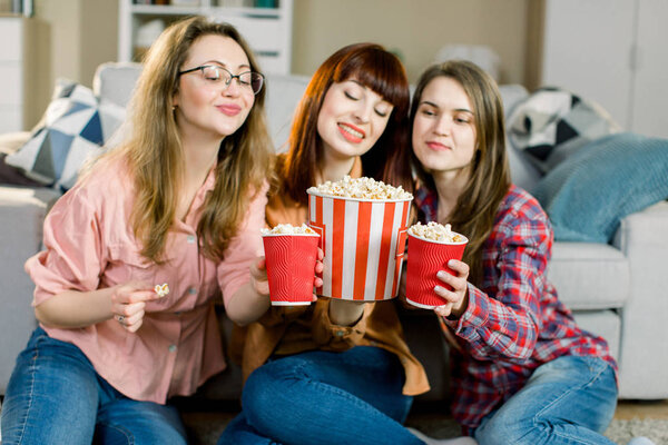 Happy three young female friends eating popcorn and sitting on the floor near sofa at home