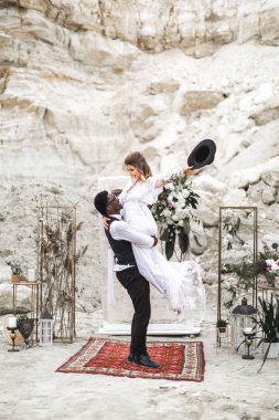 Afrikalı damat ve Kafkas gelin taze çiçeklerden düğün kemer önce bir boho tarzında birbirlerine kucaklayan. Damat gelini kollarında tutuyor, gülümsüyor ve gülüyor..