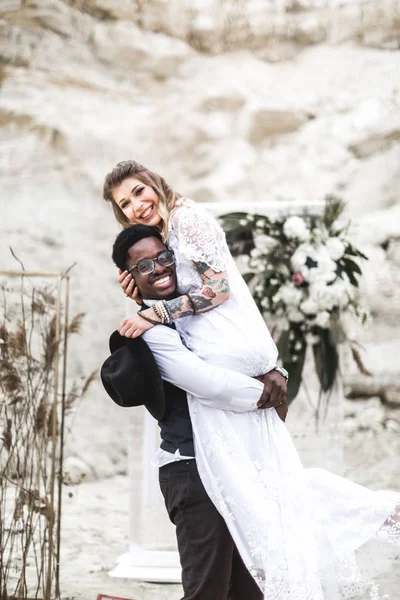 Funny hippy couple African man and Caucasian woman dressed in boho style are staying before the wedding arch in canyon outdoors, groom holds bride in arms. boho wedding. boho style.