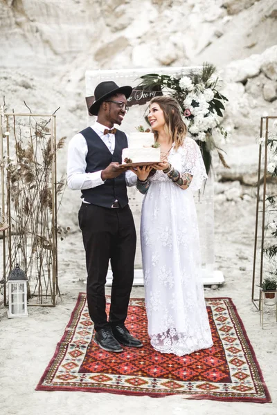 Couple African man and Caucasian woman in their boho wedding clothes holding a white cake decorated with succulents, standing on red carpet on the background of wedding arch in rustic style