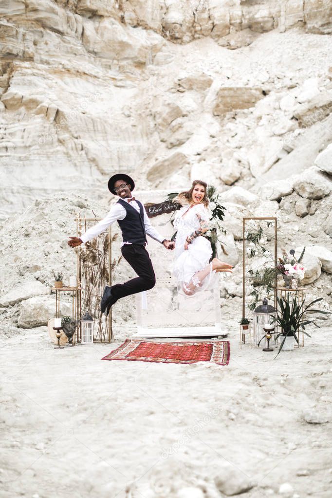 Crazy hippy multi rational Wedding couple dressed in boho style are jumping before the wedding arch in canyon outdoors. boho wedding. boho style.