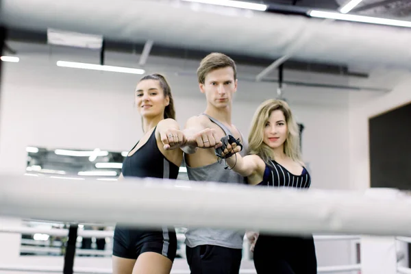 Duas meninas atraentes e belo menino estão posando no ringue de boxe dentro de casa, puxou as mãos para a frente. Fitness, conceito de boxe — Fotografia de Stock