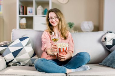 Güzel bir genç sarışın kadın kanepede oturan, şarap içme ve patlamış mısır yeme, gülümseyerek