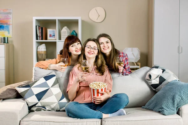 Retrato de tres alegres jóvenes amigas con palomitas de maíz, y vino, cerca del elegante sofá en casa . —  Fotos de Stock