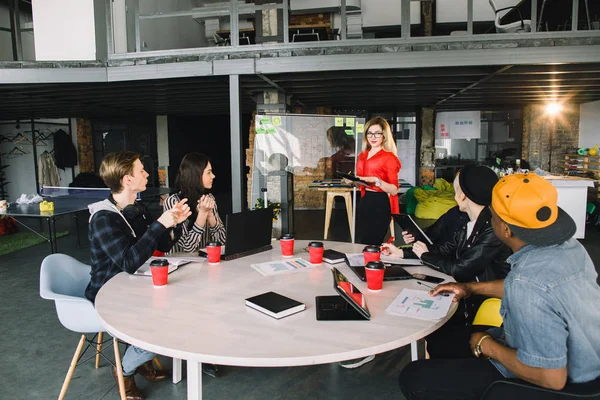 Mujer rubia joven con gafas de pie en la pared de cristal en una reunión con el equipo multinacional de trabajadores —  Fotos de Stock