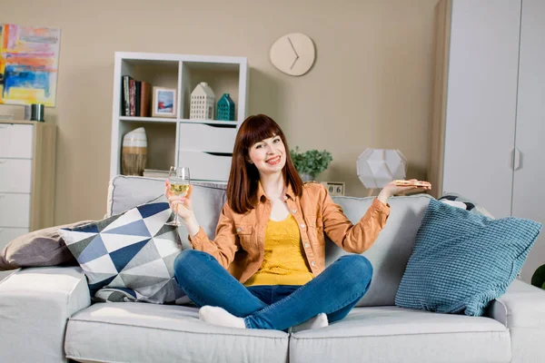 Wanita muda yang tersenyum dengan celana jeans dan kaus kuning, minum anggur dan makan pizza, sambil duduk di sofa abu-abu di rumah — Stok Foto