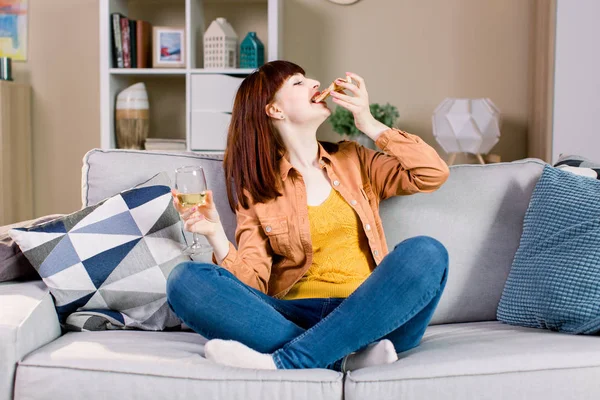 Wanita muda yang tersenyum dengan celana jeans dan kaus kuning, minum anggur dan makan pizza, sambil duduk di sofa abu-abu di rumah — Stok Foto
