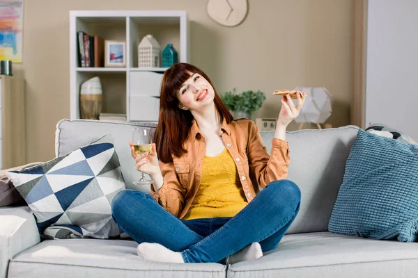 Feliz niña sonriente en jeans y camisa amarilla, sosteniendo el vaso con vino y un pedazo de pizza, mientras se sienta en el sofá gris con almohadas en casa — Foto de Stock