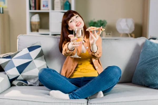 집에서 베개와 회색 소파에 앉아있는 동안, 청바지와 노란색 셔츠에 행복 한 젊은 미소 소녀, 와인과 피자 조각 유리를 들고 — 스톡 사진