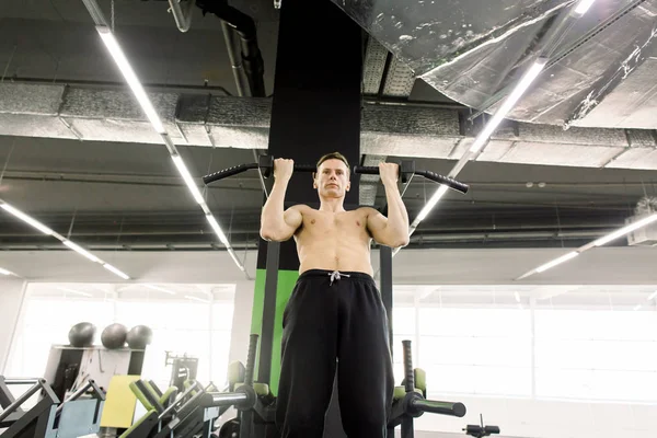 Knappe overhemdloze man doet pull-ups in de L-Sit positie op horizontale balk in de sportschool. Gymnastiek training. — Stockfoto