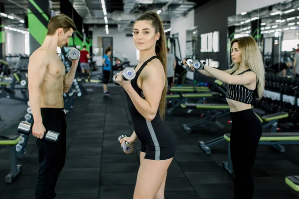 Młoda kaukaski atletyczna kobieta pompująca mięśnie z hantlami, jej przyjaciółmi, mężczyzną i kobietą na tle. Fitness dziewczyna ćwiczenia z Brzana w siłowni — Zdjęcie stockowe