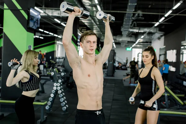 Joven guapo con el torso desnudo muscular de pie en el gimnasio y entrena bíceps con pesas junto con dos mujeres jóvenes y bonitas en el fondo —  Fotos de Stock