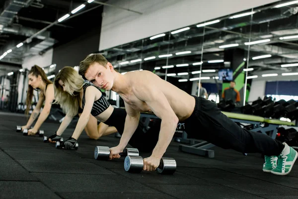 英俊的白种人和两个漂亮的女人在健身房里做着板练习, 做着沉重的哑铃 — 图库照片