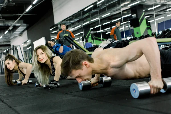 Young and sportive man couch doing push-ups with dumbbells together with two pretty women in the gym