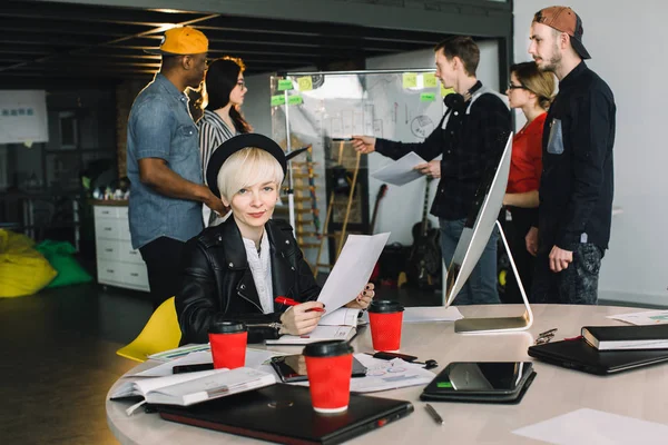 Молодая блондинка в черной шляпе и куртке сидит за столом и работает с бумагами. Женщины и мужчины случайные предприниматели, работающие над проектом стартапа возле стеклянной стены в лофт-офисе — стоковое фото