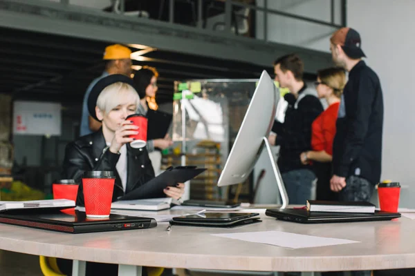 Entreprise, démarrage, planification, gestion et concept de personnes - jeune fille dans le chapeau boire du café et travailler à la table avec des ordinateurs portables et des tablettes, équipe heureuse au panneau de verre de bureau sur le fond — Photo