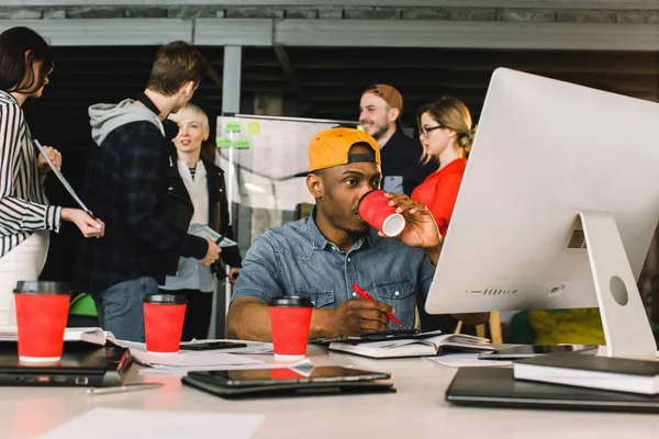 Glad afrikansk man dricker kaffe och arbetar på datorn när du sitter vid bordet i modern kontorsplats. Hans unga medarbetare står och pratar i bakgrunden. — Stockfoto