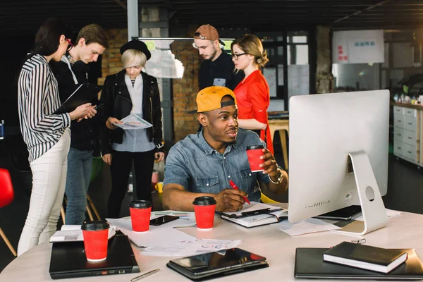テーブルに座り、近代的なロフトオフィスでコンピュータを使用しながらコーヒーを飲む若いアフリカ人男性を笑顔。ガラスの壁で話し合うカジュアルウェアの5人の若い労働者. — ストック写真