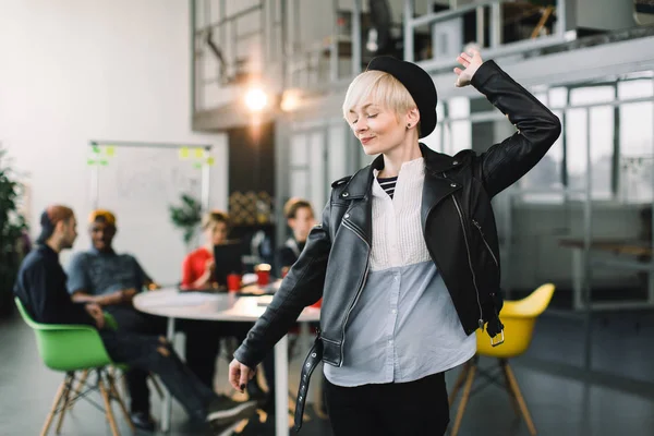 Portrait d'une employée de bureau occasionnelle debout les bras en bas, s'amusant dans un bureau lumineux, tandis que ses cinq collègues discutent à la table — Photo