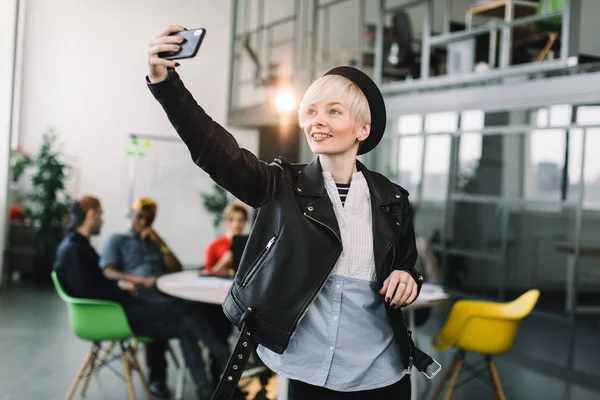 Cheerful girl in black hat and leather jacket making video call via smartphone. Young woman waving hand and smiling at phone screen. Video call concept — Stock Photo, Image