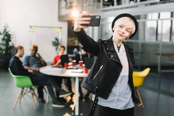 Cheerful girl in black hat and leather jacket making video call via smartphone. Young woman waving hand and smiling at phone screen. Video call concept — Stock Photo, Image