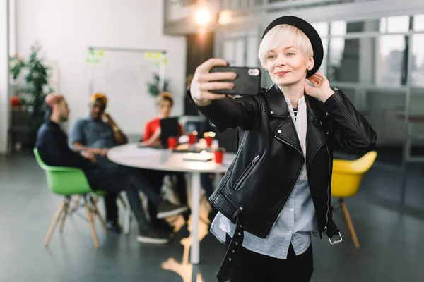 Smiling blond business woman in black leather jacket and hat making selfie on phone at small office, young workers freelancers have a meeting and discussion at the table — Stock Photo, Image