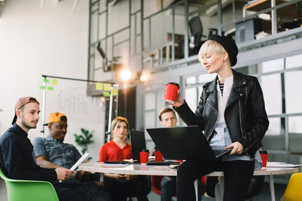 Multietnisk mångskiftande grupp av lyckliga kollegor som arbetar tillsammans i moderna Loft kontor. Kreativ ung flicka designer i hatt och skinnjacka som arbetar på laptop och sitter på bordet — Stockfoto