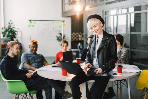 Groupe multiethnique diversifié de collègues heureux qui travaillent ensemble dans un loft moderne. Créatif jeune fille designer en chapeau et veste en cuir travaillant sur ordinateur portable et assis sur la table — Photo